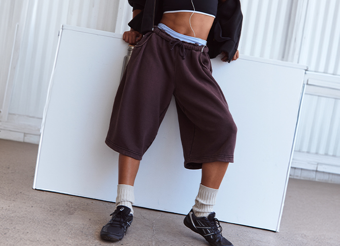 Person wearing oversized brown shorts with blue waistband, black crop top, white socks, and black sneakers in white room.