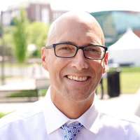 Photo of smiling man wearing glasses, a light shirt and patterned tie.