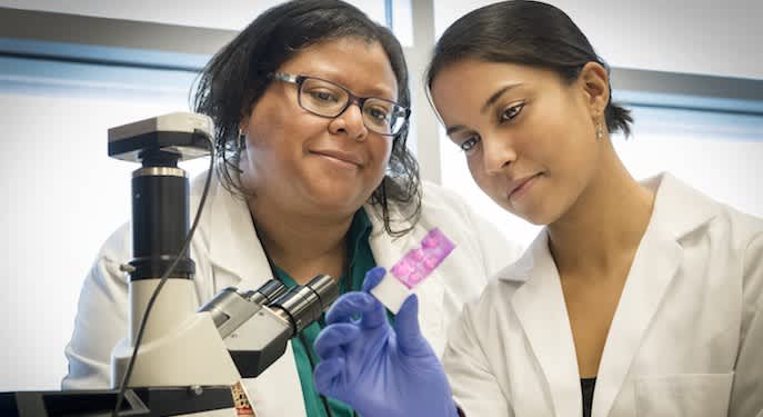 Picture of a student and professor in lab coats looking at a microscope sample. 