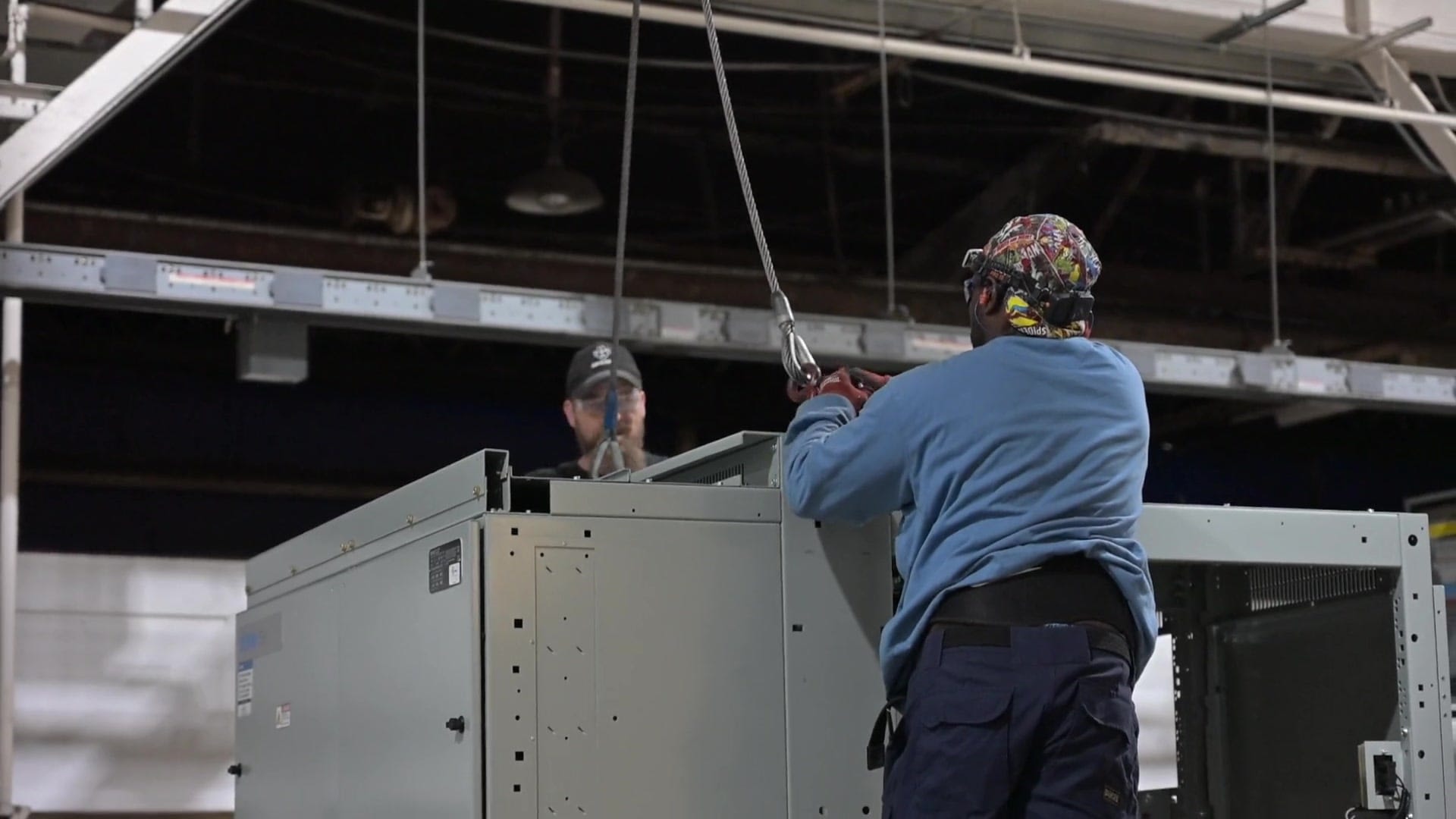 Two Beacon Modular crew members rigging a switchgear unit to an overhead hoist on the plant floor
