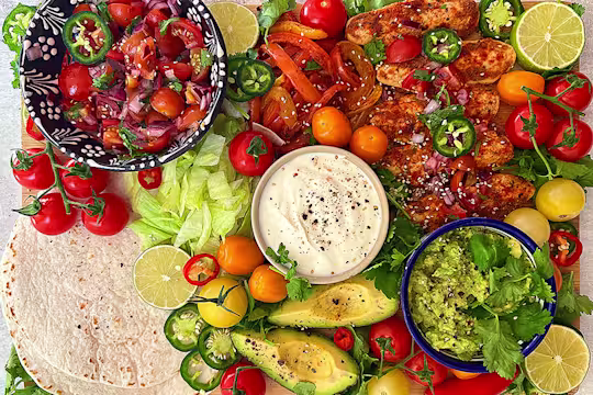Deconstructed fajita with white tortillas plus three bowls of guacamole, sour cream and fresh cherry tomatoes, salad vegetables – peppers, jalapenos, vine tomatoes avocado and cut lime.