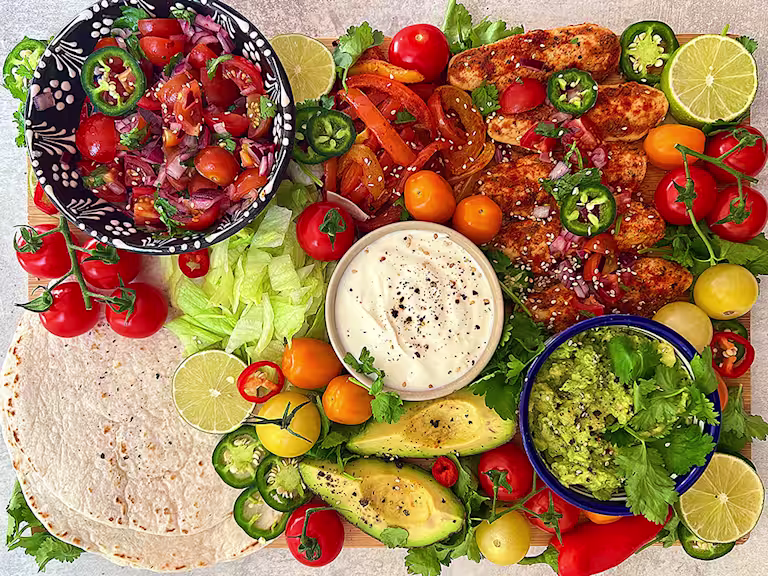 Deconstructed fajita with white tortillas plus three bowls of guacamole, sour cream and fresh cherry tomatoes, salad vegetables – peppers, jalapenos, vine tomatoes avocado and cut lime.