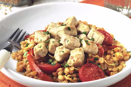 Vegetarian meal made with Quorn Pieces, pearl barley risotto, tomatoes and red pepper served in a white bowl with fork next to a glass of water.