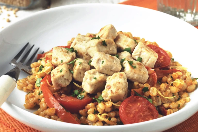 Vegetarian meal made with Quorn Pieces, pearl barley risotto, tomatoes and red pepper served in a white bowl with fork next to a glass of water.