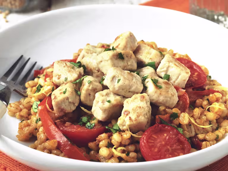 Vegetarian meal made with Quorn Pieces, pearl barley risotto, tomatoes and red pepper served in a white bowl with fork next to a glass of water.