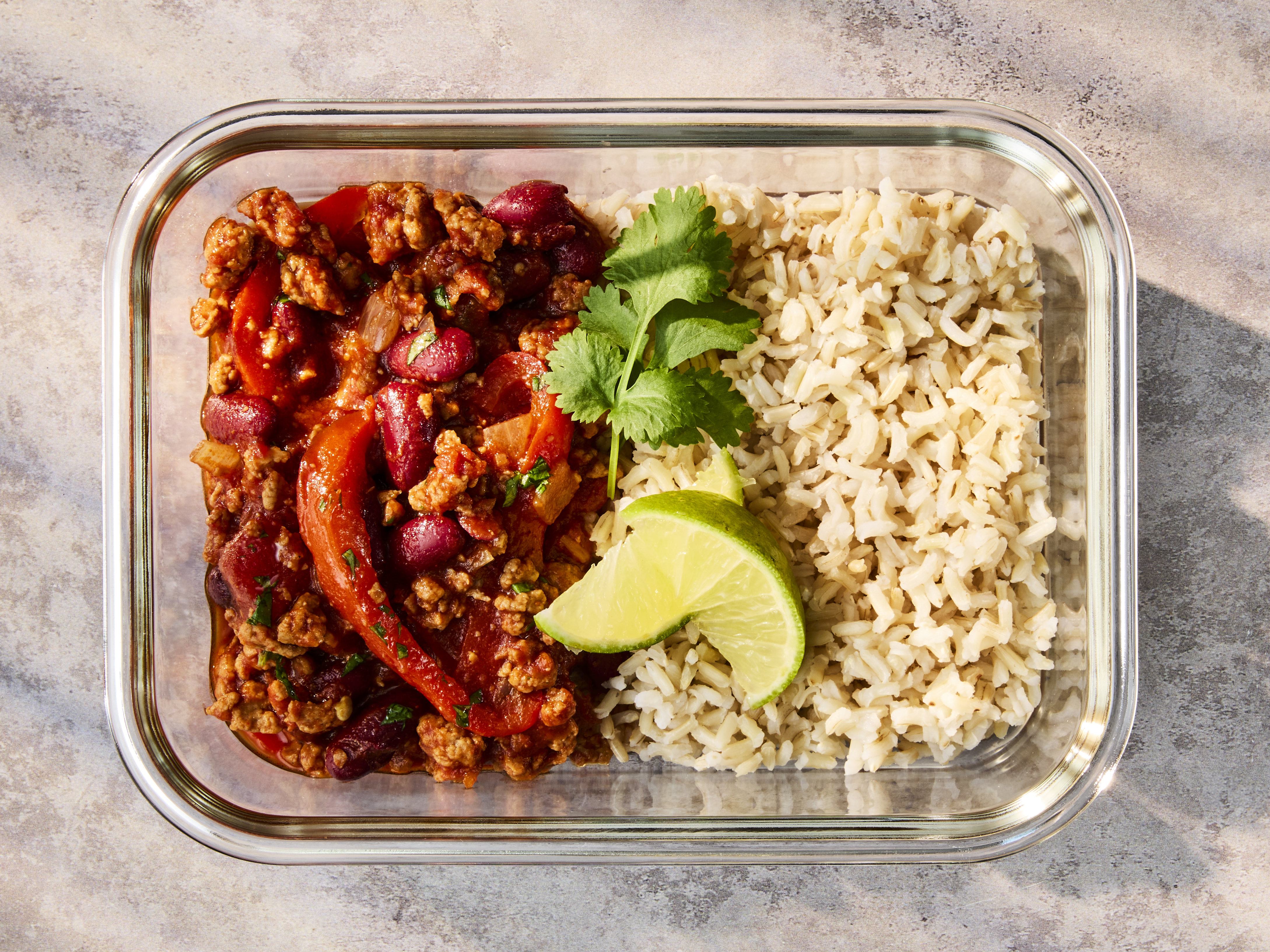 A glass meal prep container holding Quorn Mince Chilli with Rice, garnished with lime and herbs.