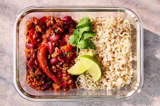 A glass meal prep container holding Quorn Mince Chilli with Rice, garnished with lime and herbs.
