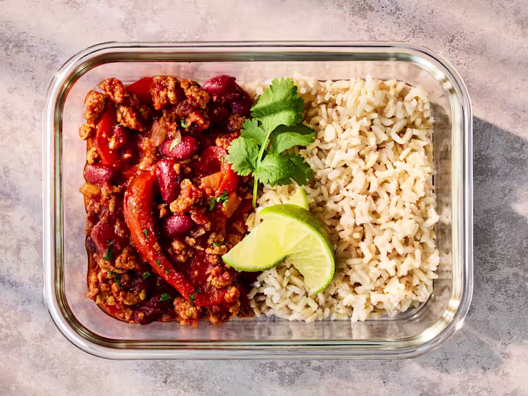 A glass meal prep container holding Quorn Mince Chilli with Rice, garnished with lime and herbs.