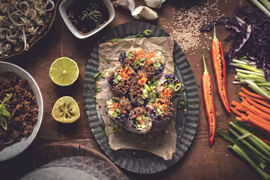 Spring rolls made with cucumber, red cabbage, carrot, bean sprouts, and Quorn Grounds arranged on a plate surrounded by lime halves, chiles, sesame seeds, and their components with a sauce on the side.