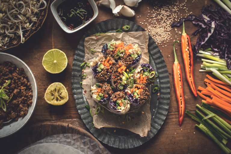 Spring rolls made with cucumber, red cabbage, carrot, bean sprouts, and Quorn Grounds arranged on a plate surrounded by lime halves, chiles, sesame seeds, and their components with a sauce on the side.