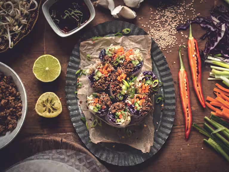 Spring rolls made with cucumber, red cabbage, carrot, bean sprouts, and Quorn Grounds arranged on a plate surrounded by lime halves, chiles, sesame seeds, and their components with a sauce on the side.