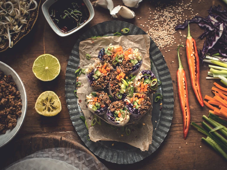 Spring rolls made with cucumber, red cabbage, carrot, bean sprouts, and Quorn Grounds arranged on a plate surrounded by lime halves, chiles, sesame seeds, and their components with a sauce on the side.