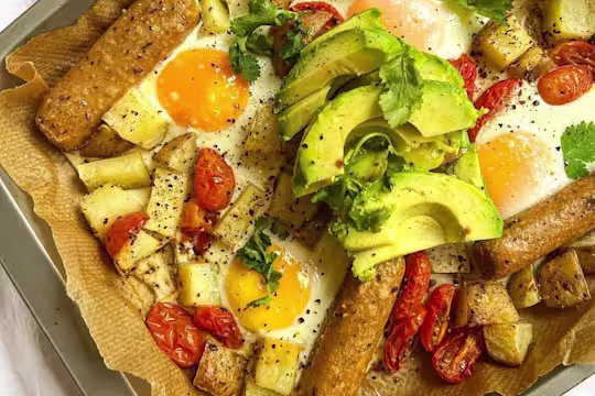 Baking tray lined with baking paper with Quorn vegetarian Sausages, potatoes, tomato and runny eggs garnished with avocado and fresh coriander.