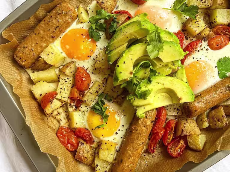 Baking tray lined with baking paper with Quorn vegetarian Sausages, potatoes, tomato and runny eggs garnished with avocado and fresh coriander.