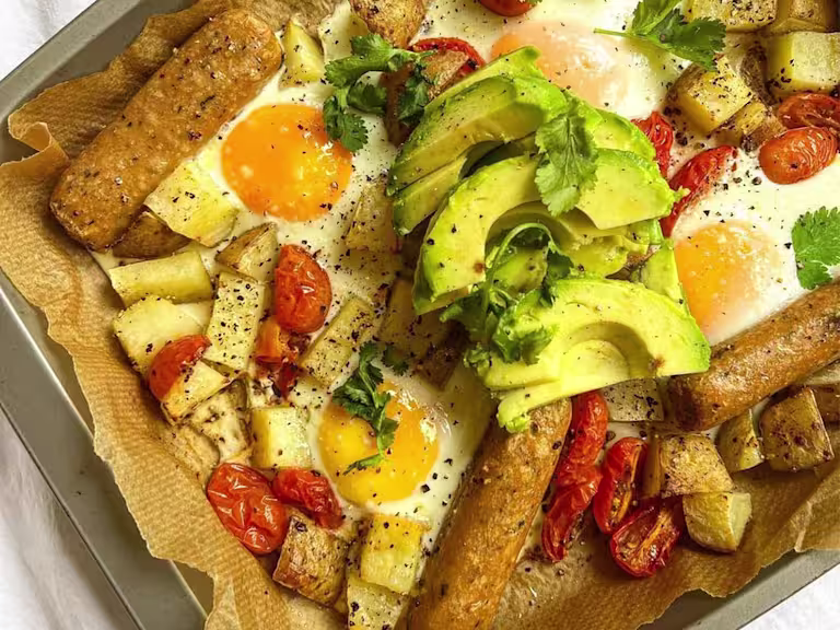 Baking tray lined with baking paper with Quorn vegetarian Sausages, potatoes, tomato and runny eggs garnished with avocado and fresh coriander.