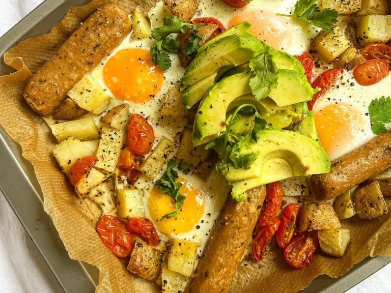 Baking tray lined with baking paper with Quorn vegetarian Sausages, potatoes, tomato and runny eggs garnished with avocado and fresh coriander.