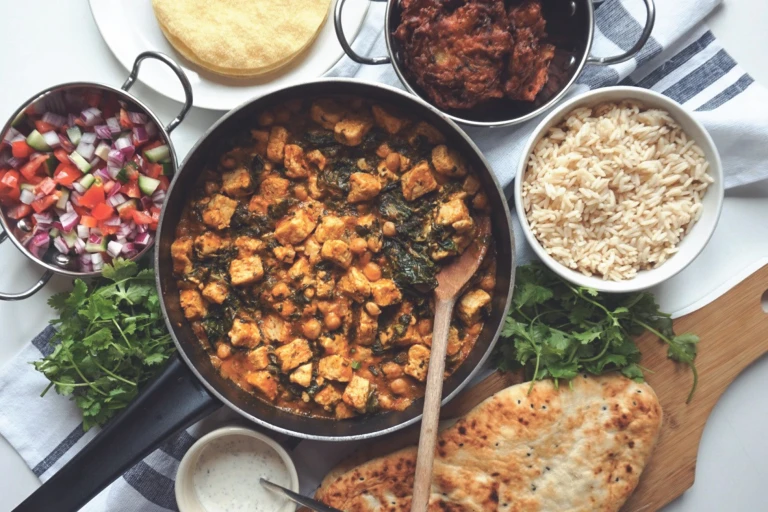A curry made with Quorn Pieces, chickpeas, and spinach in a large pot surrounded by a bowl of rice, a bowl of salad, a plate of poppadoms, and naan bread on a board.