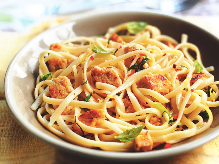 A taupe bowl of linguine topped with Quorn Pieces, chili flakes, and basil on an ochre tablecloth.