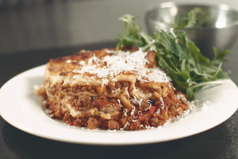 A portion of vegetarian lasagne made with Quorn Mince topped with Parmesan cheese served with a side of rocket in a white bowl