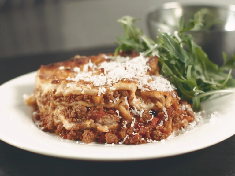 A portion of vegetarian lasagne made with Quorn Mince topped with Parmesan cheese served with a side of rocket in a white bowl