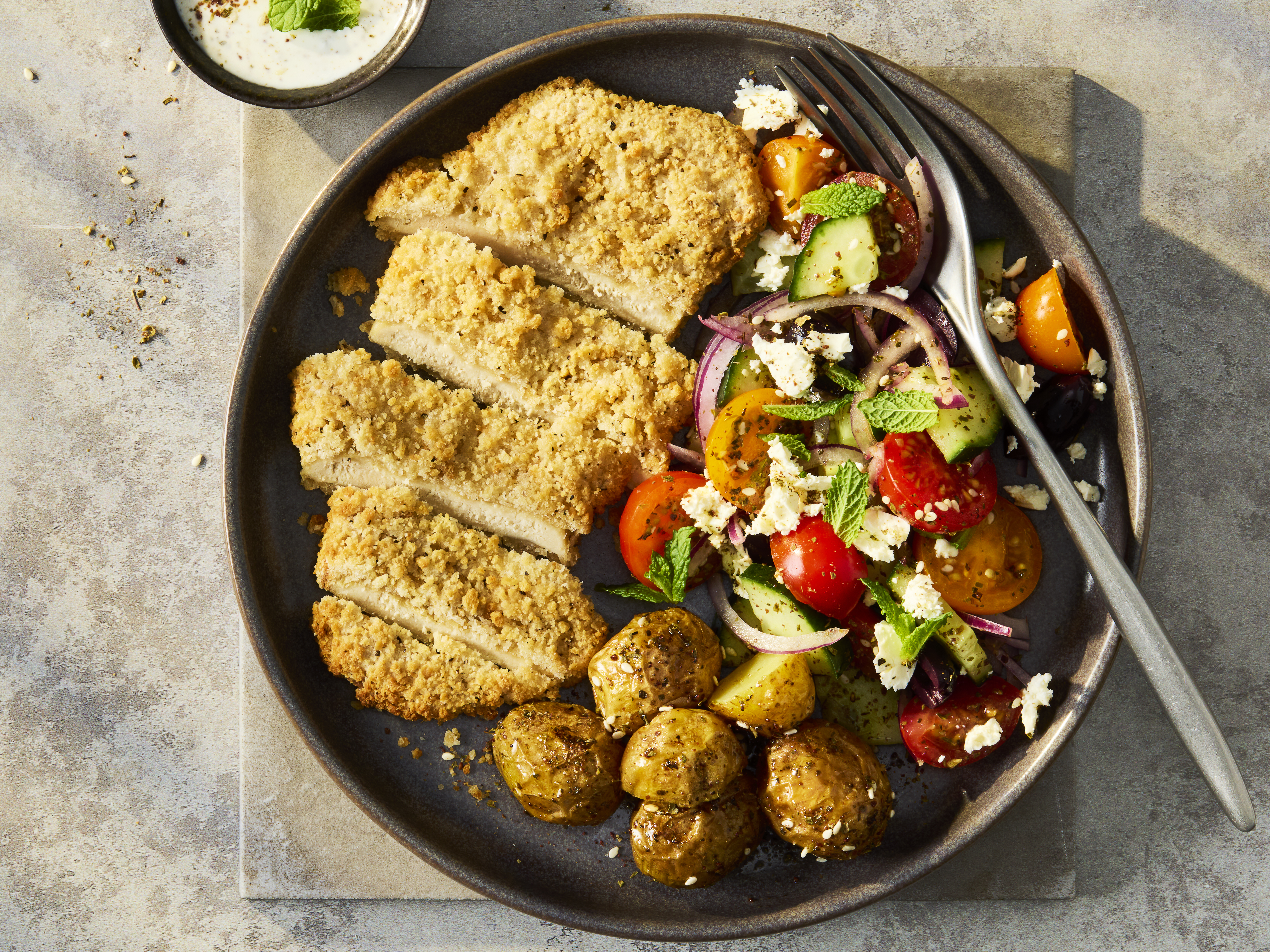 A vibrant Greek salad featuring crisp cucumbers, cherry tomatoes, Kalamata olives, and crumbled feta, topped with a sliced Quorn Lemon and Pepper Schnitzel and cripsy potatoes.