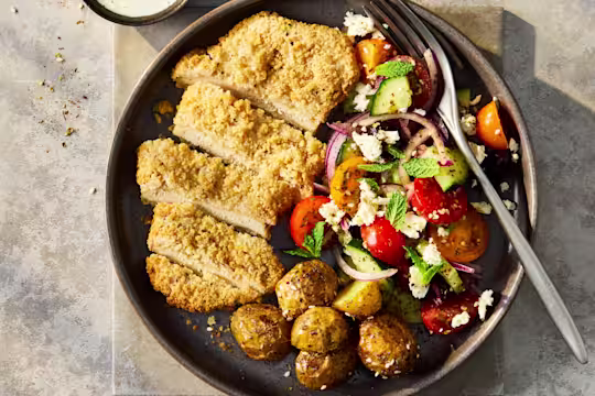 A vibrant Greek salad featuring crisp cucumbers, cherry tomatoes, Kalamata olives, and crumbled feta, topped with a sliced Quorn Lemon and Pepper Schnitzel and cripsy potatoes.