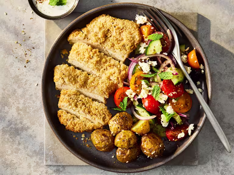 A vibrant Greek salad featuring crisp cucumbers, cherry tomatoes, Kalamata olives, and crumbled feta, topped with a sliced Quorn Lemon and Pepper Schnitzel and cripsy potatoes.