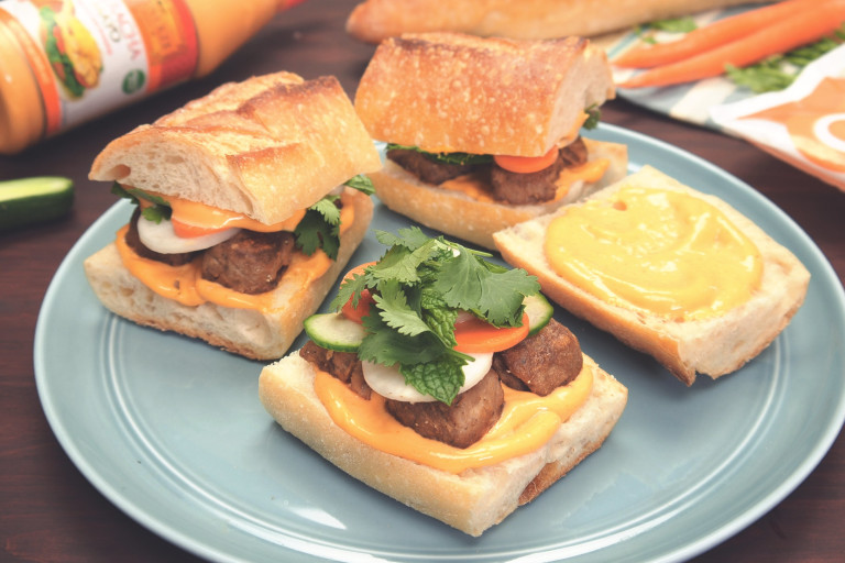 Two small banh mi sandwiches with pickled carrots and daikon, cucumber, Quorn Meatballs, cilantro, mint, and sriracha mayo on a baguette atop a blue plate.
