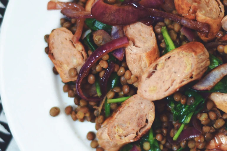Lentils topped with caramelised red onions, spinach, and Quorn Sausages on a white plate.