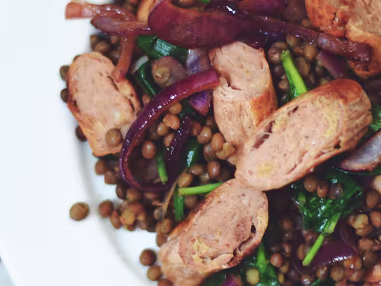 Lentils topped with caramelised red onions, spinach, and Quorn Sausages on a white plate.