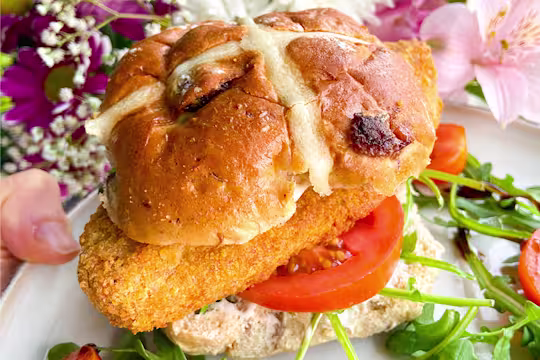 A Quorn Crunchy Fillet Burger in a hot cross bun on a plate with flowers in the background.