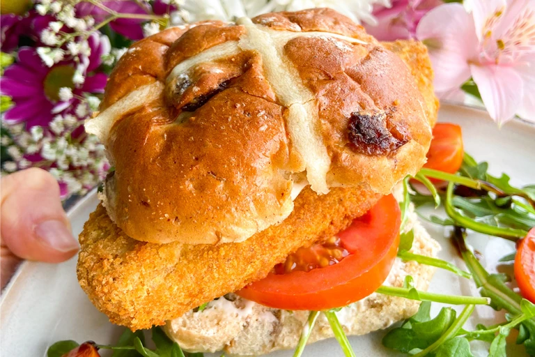 A Quorn Crunchy Fillet Burger in a hot cross bun on a plate with flowers in the background. 