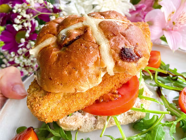 A Quorn Crunchy Fillet Burger in a hot cross bun on a plate with flowers in the background.