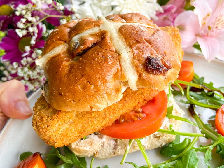 A Quorn Crunchy Fillet Burger in a hot cross bun on a plate with flowers in the background. 