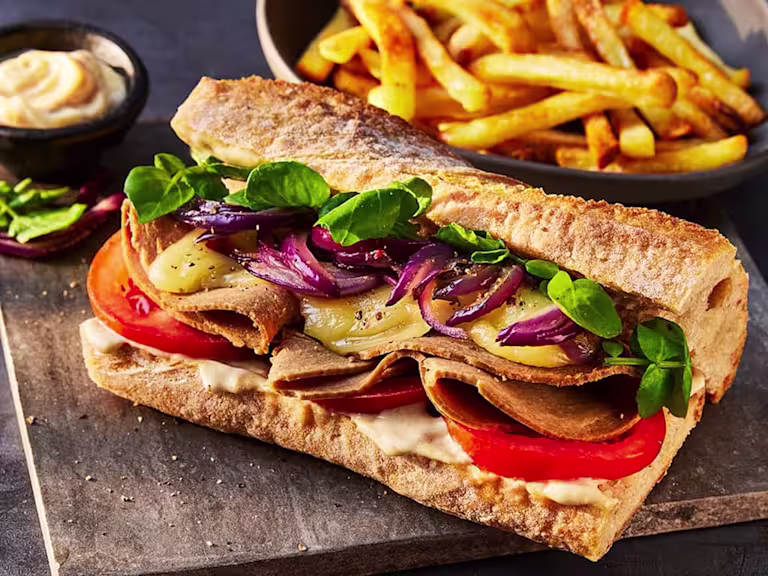 A Quorn Vegetarian Roast Beef Sandwich with melted cheese, caramelised onions, watercress, tomatoes and fries on a wooden background.