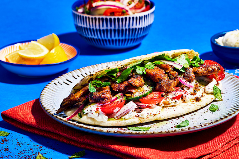 A pitta topped with Quorn Makes Amazing Turkish Style Kebab, tomatoes, cucumber, red onion, yoghurt sauce, and herbs on a red placemat on a cobalt blue background.