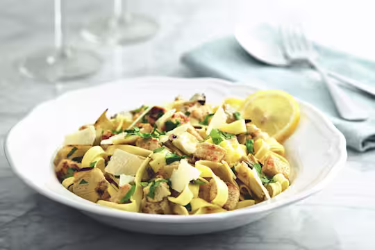 A white bowl full of tagliatelle pasta topped with Quorn Pieces, artichokes, parsley and parmesan, garnished with a lemon slice against a background of two wine glasses and a fork and spoon on a light blue napkin.