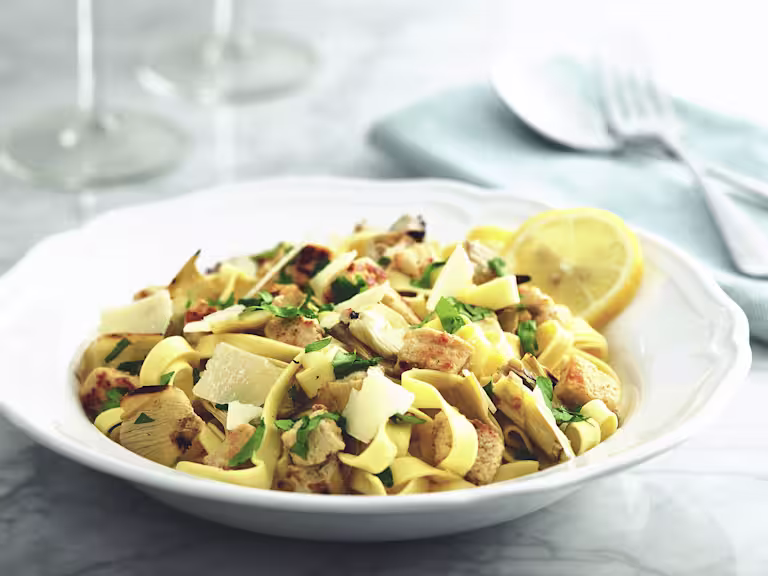 A white bowl full of tagliatelle pasta topped with Quorn Pieces, artichokes, parsley and parmesan, garnished with a lemon slice against a background of two wine glasses and a fork and spoon on a light blue napkin.