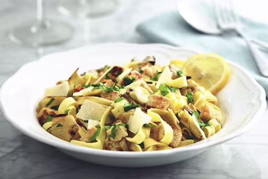Tagliatelle pasta with Quorn Pieces and artichokes topped with cheese and parsley with a lemon wedge on the side.