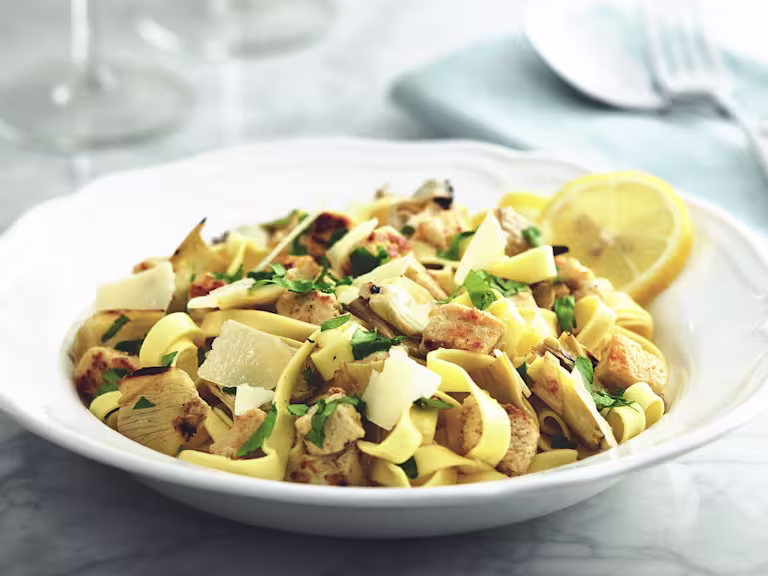 Tagliatelle pasta with Quorn Pieces and artichokes topped with cheese and parsley with a lemon wedge on the side.
