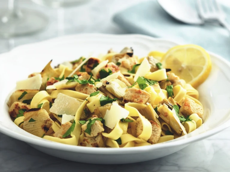 Tagliatelle pasta with Quorn Pieces and artichokes topped with cheese and parsley with a lemon wedge on the side.