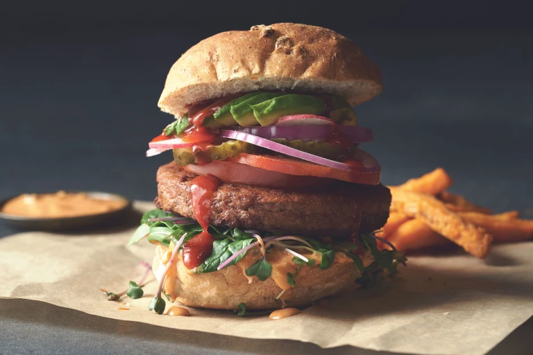 A Quorn Quarter Pounder piled high with burger sauce, greens, tomato slices, gherkins, radishes, red onion, and avocado.