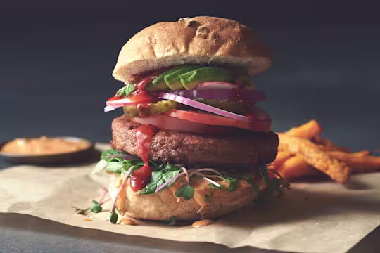 A Quorn Quarter Pounder piled high with burger sauce, greens, tomato slices, gherkins, radishes, red onion, and avocado.