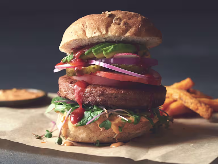 A Quorn Quarter Pounder piled high with burger sauce, greens, tomato slices, gherkins, radishes, red onion, and avocado.