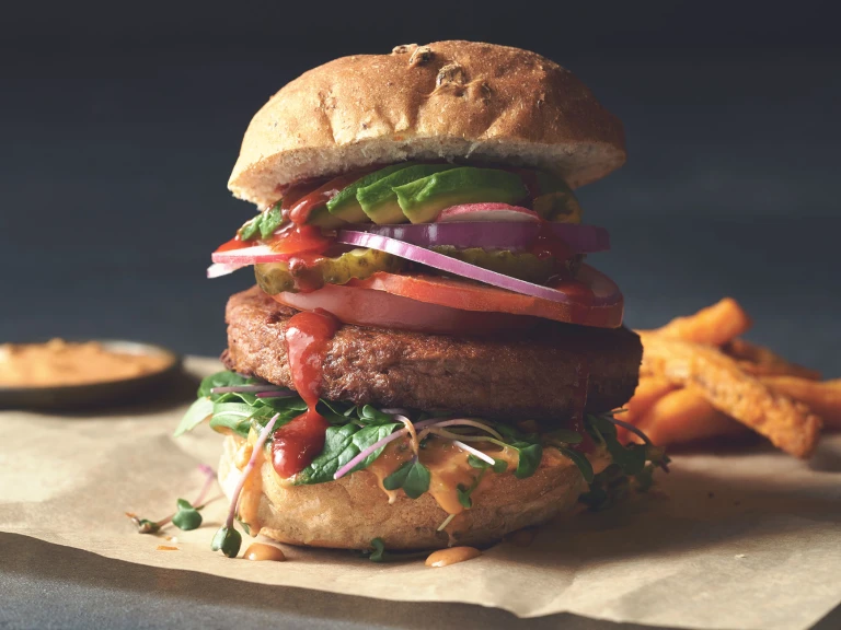 A Quorn Quarter Pounder piled high with burger sauce, greens, tomato slices, gherkins, radishes, red onion, and avocado.