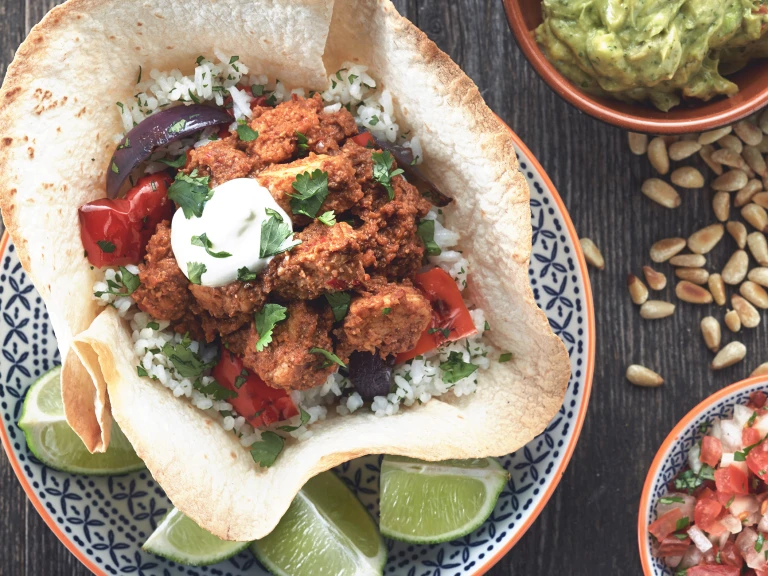 A tortilla bowl filled with cilantro rice, grilled vegetables, and Quorn Pieces, topped with sour cream with lime wedges on the side.