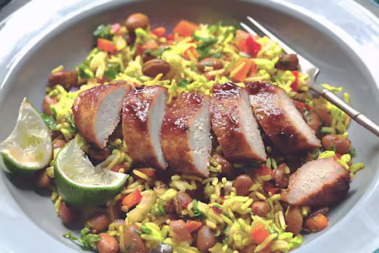 A sliced Quorn Fillet topped with glaze atop a bed of rice with beans and peppers with two wedges of lime on the side.