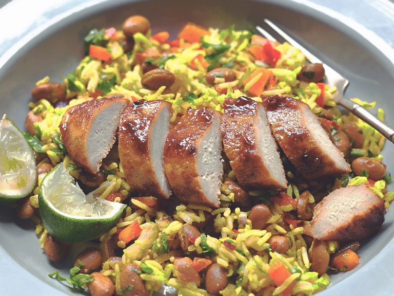 A sliced Quorn Fillet topped with glaze atop a bed of rice with beans and peppers with two wedges of lime on the side.