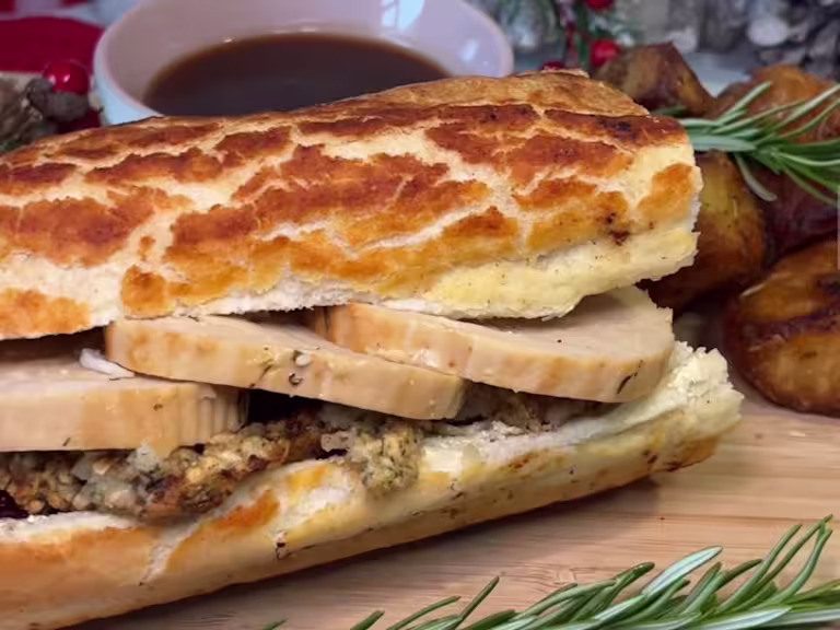 A Quorn Christmas Roast & Stuffing Baguette on a wooden board with a bowl of gravy.