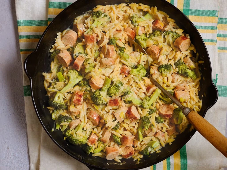 A large portion of Quorn Vegetarian Sausage and Broccoli Orzo served in a dark dish with a serving spoon.